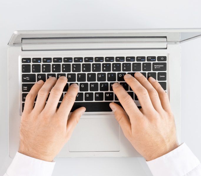 Flat layout of hands of nurse with pen filling in medical history form while sitting by desk next to male colleague using laptop at workplace